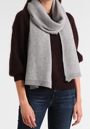 Femme portant une écharpe tricotée texturée gris clair sur un pull bordeaux foncé et un jean bleu, debout devant un fond uni.