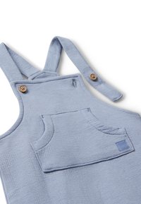 Salopette en coton bleu clair avec des bretelles réglables, des boutons en bois et une poche frontale. Tissu texturé avec un motif tissé.