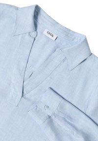 Camicia azzurro chiaro con colletto e scollo a V, dotata di un polsino a bottone singolo, realizzata in tessuto liscio. Presenta un'etichetta con il marchio "CECIL".