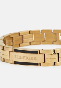 Zlatna metalna narukvica s pravokutnim karikama, s crnim akcentom i ugraviranim detaljem "HILFIGER" na središnjoj pločici.