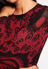 Top a fantasia rossa e nera con un design a maglia testurizzata caratterizzato da motivi circolari e astratti; scollatura rotonda, maniche lunghe.