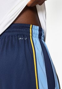 Calções atléticos em azul marinho e azul claro com uma textura respirável, apresentando riscas laterais amarelas e um cós elástico. Logótipo "DRY-FIT" visível.