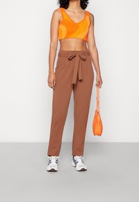 Bruine high-waisted broek met een strikdetail in de taille, gecombineerd met een oranje patroon crop top en witte sneakers.