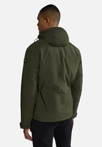 Giacca a vento verde oliva con texture liscia, coulisse regolabile e polsini elastici; presenta un'etichetta sulla spalla sinistra.