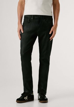 Homme portant un jean noir ajusté, des baskets noires avec des rayures blanches, et un t-shirt blanc, debout devant un fond clair uni.