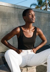 Femeie într-un top negru și pantaloni albi stă afară pe trepte de piatră, cu mâinile în buzunare, uitându-se cu încredere în depărtare, sub lumina soarelui.