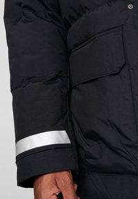 Black puffer jacket sleeve with silver reflective stripe, textured fabric, and a visible patch pocket. "Spray Tech" logo partially visible.