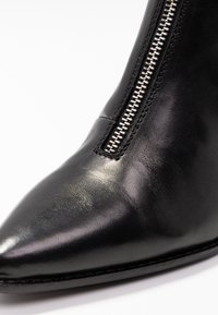 Acercamiento de una bota de cuero negra con punta afilada y un zipper plateado en el lado sobre un fondo blanco.