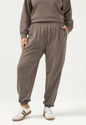 Persoon draagt taupe joggerbroek met elastische boorden en tailleband, gecombineerd met witte sneakers met zwarte strepen, staand met één hand in de zak.