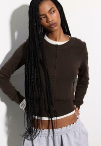 Brauner Cardigan mit Knopfleiste, rundem Ausschnitt, gerippter Struktur und weißen Akzenten an den Bündchen und am Saum, kombiniert mit hellgrauen Hosen.