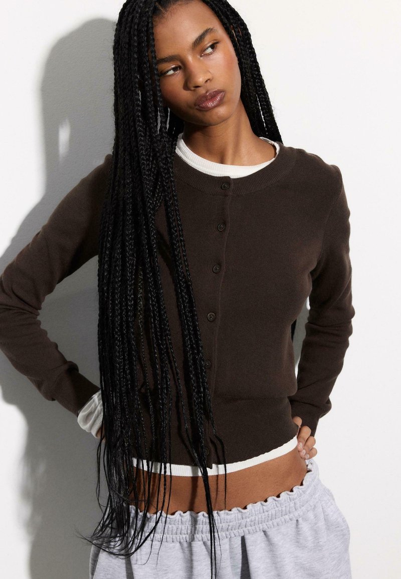 Brauner Cardigan mit Knopfleiste, rundem Ausschnitt, gerippter Struktur und weißen Akzenten an den Bündchen und am Saum, kombiniert mit hellgrauen Hosen.