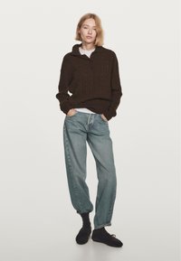 Donna in piedi con le mani in tasca, indossa un maglione marrone scuro, jeans azzurri chiari, calzini neri e scarpe stringate nere su uno sfondo bianco.