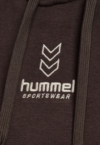 Nahaufnahme eines dunkelbraunen Sportbekleidungsstoffs mit weißem gesticktem Hummel-Logo und Chevron-Symbolen zwischen den Zugbändern.