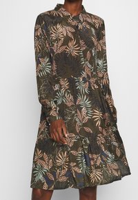 Langärmliges Kleid in Dunkelgrün mit einem Blättermuster in Pink und Blau. Mit kragen und durch Knöpfe geschlossenen Ausschnitt, sowie einem ausgestellten, gestuften Saum. Leichtes Material.