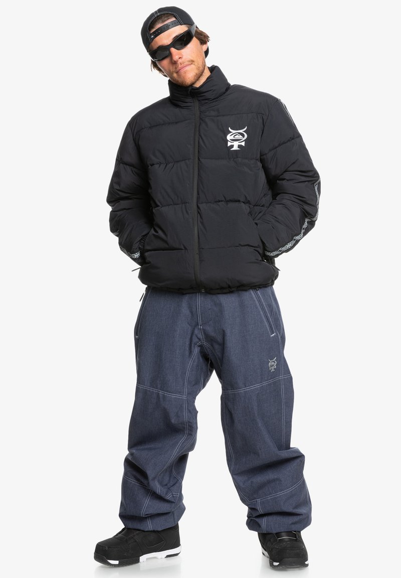 Czarna kurtka puchowa z zamkiem błyskawicznym z przodu i logo na lewej piersi, w połączeniu z luźnymi niebieskimi dżinsami i czarnymi butami. Minimalistyczny design.