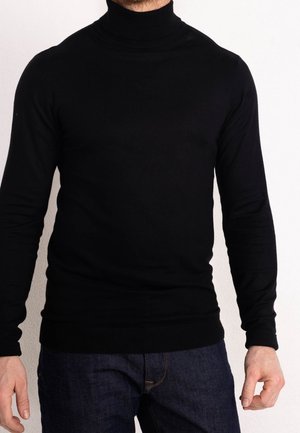 Hombre con un suéter de cuello alto negro ajustado y jeans azul oscuro, de pie contra un fondo blanco liso.