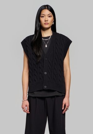 Femme aux longs cheveux noirs portant un gilet sans manches en maille torsadée noire, une chemise noire et un pantalon noir sur un fond gris.