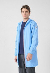 Hellblaue wasserdichte Jacke mit Kapuze, Frontreißverschluss, elastischen Bündchen und kontrastierenden roten Akzenten, kombiniert mit einem dunklen Hemd und grauen Hosen.