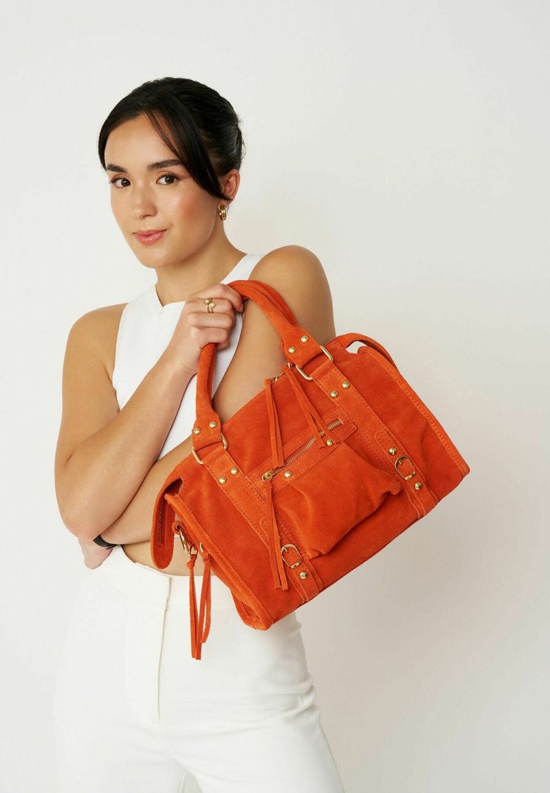 Sac en daim orange avec quincaillerie dorée, à double poignées, poche zippée à l'avant et accents de pampilles, tenu devant une personne portant un haut blanc.