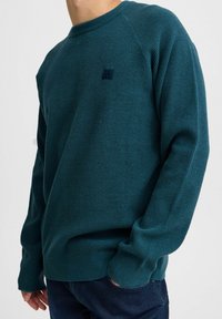 Teal bordázott pulóver kerek nyakkal, hosszú ujjal és kis logóval a mellkason, sötét farmernadrággal párosítva.