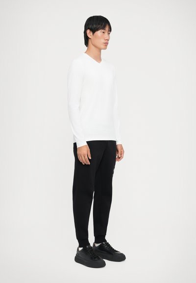 Un top alb cu mânecă lungă și decolteu în V, asortat cu pantaloni joggeri negri și adidași negri, evidențiind un design casual și o siluetă ajustată.