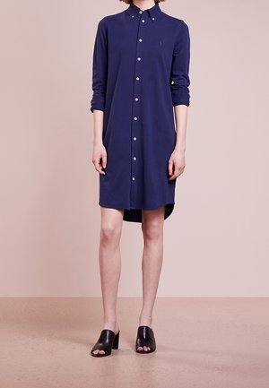Navyblå skjortekjole med knapper og lange ærmer, lavet af blødt stof. Har krave og sideslids, kombineret med sorte mule-sandaler med åben tå.