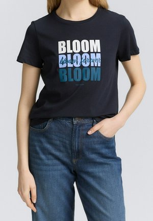 Femme portant un t-shirt noir avec le texte "BLOOM bloom forever BLOOM" en blanc et bleu, associé à un jean bleu, main dans la poche.