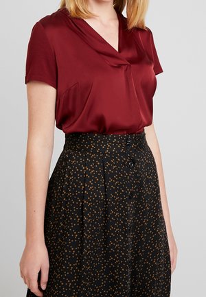 Kobieta w bordowej bluzce z krótkim rękawem, włożonej w wysoką czarną spódnicę w brązowe groszki z guzikami z przodu.