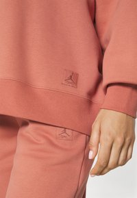 Completo con felpa a maniche lunghe e pantaloni in tonalità corallo. Tessuto morbido con texture liscia. Presenta loghi Jordan in rilievo su entrambi i capi.