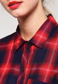 Rood en marineblauw geruite shirt met een button-down kraag, gemaakt van een gestructureerde stof en met zichtbare knopen op de voorkant.