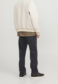 Krémová bomber bunda s béžovými pruhovanými manžetami přes hnědou vrstvenou košili, doplněná tmavě šedými džínami a černými teniskami.