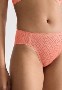 Close-up van een persoon die koraalkleurige hoge taille ondergoed en een bijpassende bh draagt, met zichtbare textuur van de stof en contrast met de huidtoon.