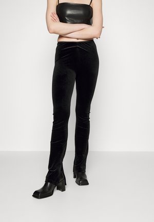 Personne portant un crop top en cuir noir, un pantalon ajusté en velours noir et des bottes noires à talons, debout les bras croisés.