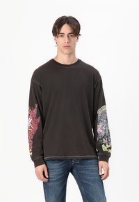 Černé tričko s dlouhými rukávy a barevnými grafickými vzory na rukávech; má drsnou texturu a viditelné švy. Kombinováno s modrými džínami.