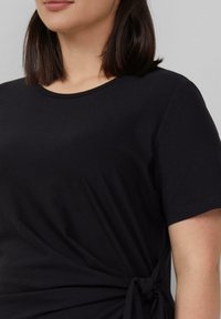 Schwarzes T-Shirt mit rundem Halsausschnitt und kurzen Ärmeln, das an der Seite mit einem Knoten-Detail für einen gerafften Effekt versehen ist. Weicher Stoff.