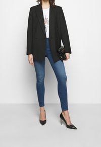 Blazer negro de doble botonadura, camiseta gráfica blanca ajustada, jeans ajustados azules y tacones de aguja negros, sosteniendo un pequeño clutch negro.