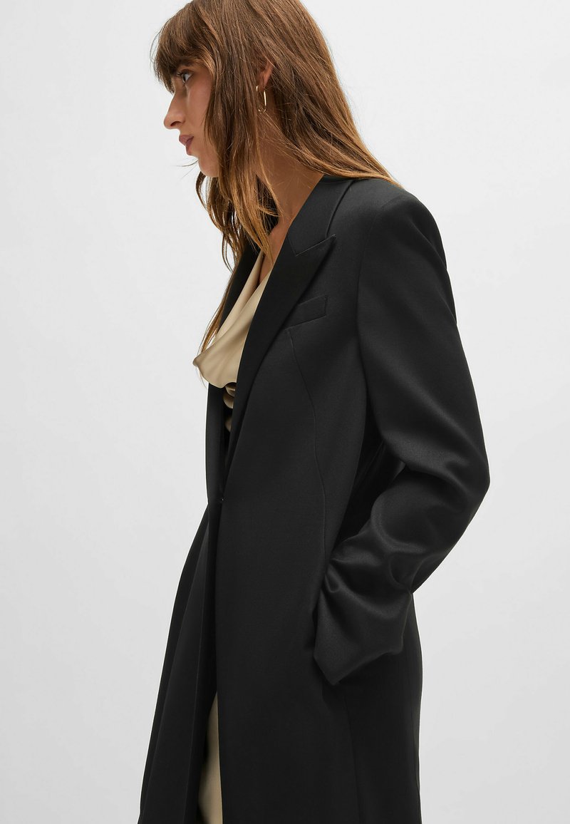 Blazer noir sur mesure avec une texture lisse, des revers crantés et deux poches latérales, porté sur un haut crème. Modèle vu de profil.