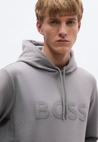 Hall kapuutsiga pusa, mis on valmistatud pehmest kangast ja millel on rinnal esile tõstetud reljeefne "BOSS" logo ning nööriga reguleeritav kapuuts.