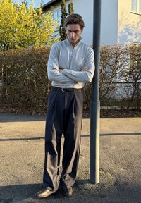 Mladý muž so skríženými rukami, oblečený v sivom pulóvri a tmavých nohaviciach, stojaci vedľa pouličného lampu počas slnečného dňa vonku.