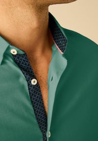 Cămașă verde închis cu guler navy cu model, având nasturi albi și accente subtile de roz. Textură netedă, design modern.