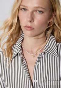 Camicia a righe verdi e bianche con colletto e taschino frontale, indossata con una collana di perle con un ciondolo d'oro.