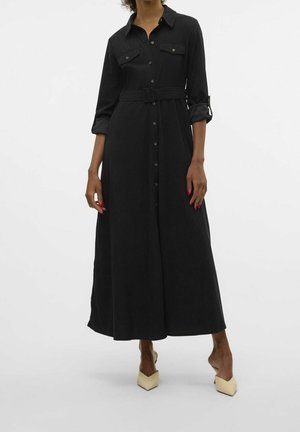 Femme portant une robe noire à manches longues boutonnée avec une ceinture, debout pieds nus sur un fond clair uni.