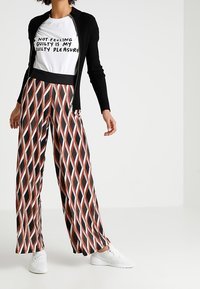 Gilet noir à côtes porté sur un t-shirt graphique blanc ; pantalon large avec un motif géométrique de diamants noirs, rouges et roses ; baskets blanches.