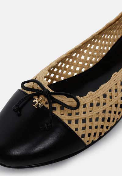 Chaussure plate en cuir noir avec des côtés beiges tissés et un nœud noir orné d’un petit charm décoratif doré.