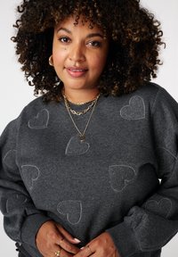 Vrouw met krullend haar draagt een donkergrijze trui met geborduurde hartpatronen, gouden ringoorbellen, gelaagde gouden kettingen en ringen.