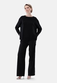 Schwarzer übergroßer Pullover mit Rundhalsausschnitt und gesenkten Schultern, kombiniert mit maßgeschneiderten schwarzen Hosen und silbernen Absatzsandalen.