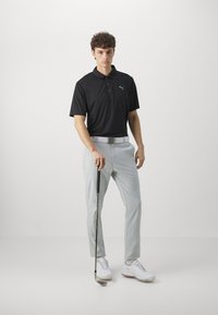 Crna polo majica s kratkim rukavima i tri gumba, svijetlosive hlače i bijele golf cipele. Drži golf palicu.