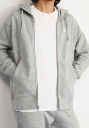 Osoba ubrana w jasnoszary, rozpinany na zamek kaptur Nike na białej koszulce, do tego pasujące szare spodnie dresowe, ręce w kieszeniach bluzy.