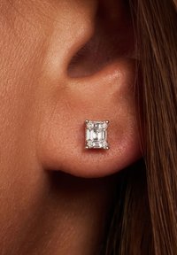 Vierkante diamanten studoorbel met een zilveren setting, met een centrale smaragdgeslepen diamant omringd door kleinere diamanten. Achtergrond is een huidskleur.