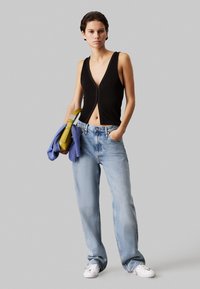 Top nero senza maniche con scollatura a V profonda e zip frontale, abbinato a jeans a gamba larga blu chiaro. Tiene un maglione viola e una borsa gialla.